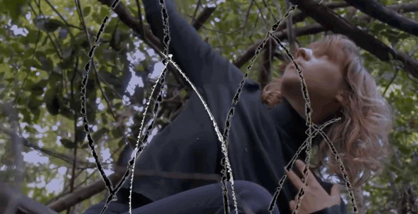 A young woman with a black turtleneck shirt sits on tree branches, looking up. 
